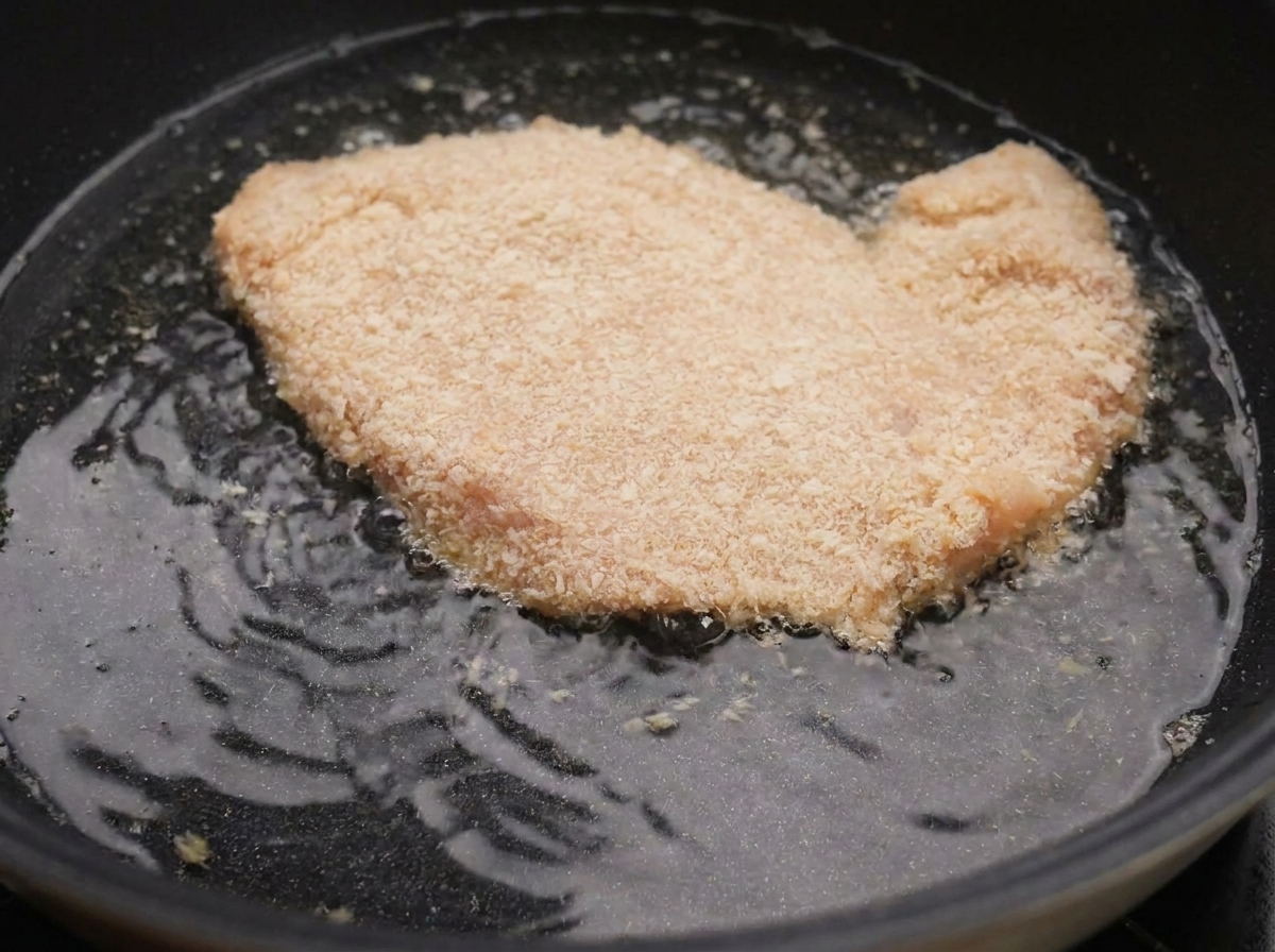Ein paniertes Hähnchenschnitzel brutzelt in heißem Öl in einer schwarzen Pfanne.