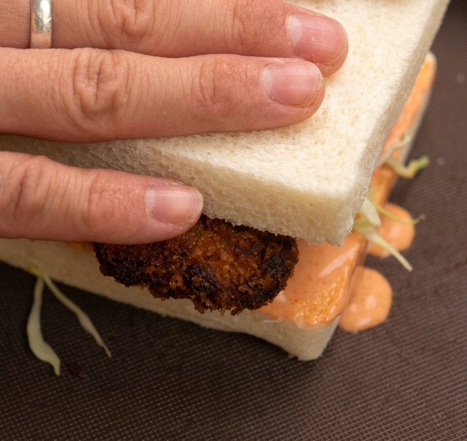Hände drücken die obere Brotscheibe auf das fertige Chicken Sandwich.