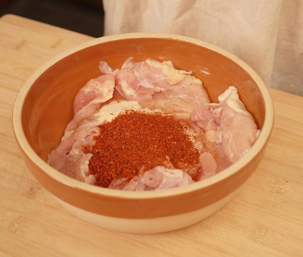 Rohe Hähnchenstücke in einer braunen Schüssel mit rotem Chilipulver und Gewürzen.
