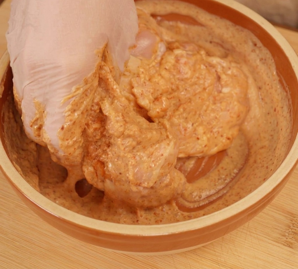 Eine behandschuhte Hand vermengt rohe Hähnchenstücke gründlich in einer cremigen, orangefarbenen Joghurt-Marinade.