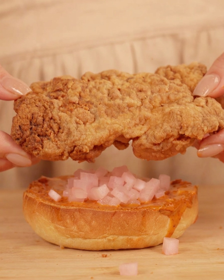 Hände platzieren ein Hähnchenfilet auf ein Brötchen mit Sauce und rosa Rettichwürfeln.