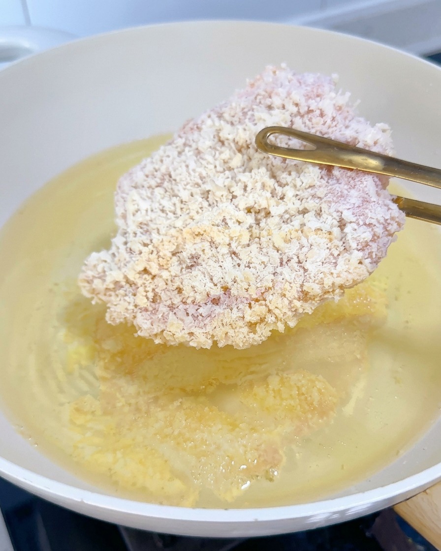 Ein gefrorenes, paniertes Hähnchenschnitzel wird mit einer goldenen Zange in sprudelndes heißes Öl gleiten gelassen.