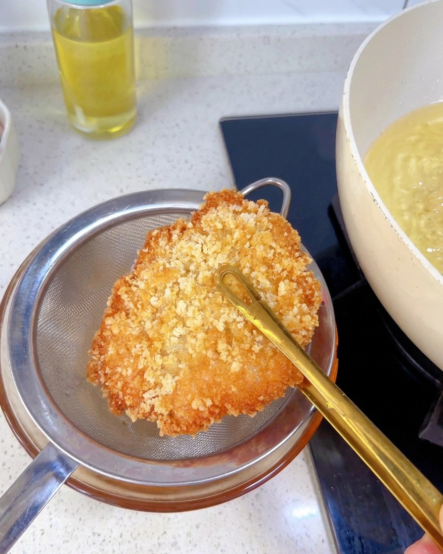 Ein fertig frittiertes, goldbraunes Hähnchenschnitzel wird mit einer Zange über einem Metallsieb gehalten, um überschüssiges Öl abtropfen zu lassen.
