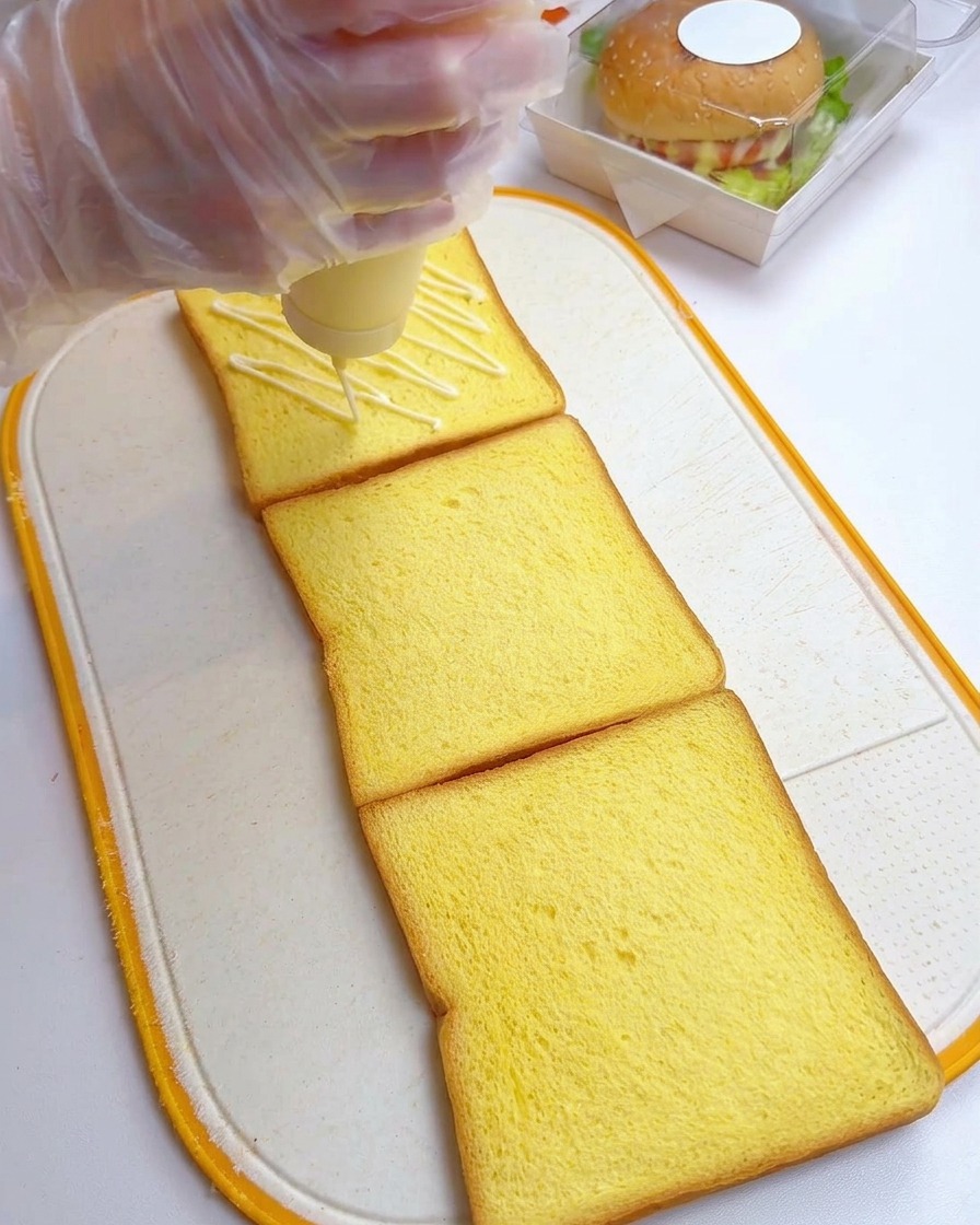 Eine Hand mit Handschuh verteilt Mayonnaise im Zickzack-Muster auf drei Scheiben Toastbrot.