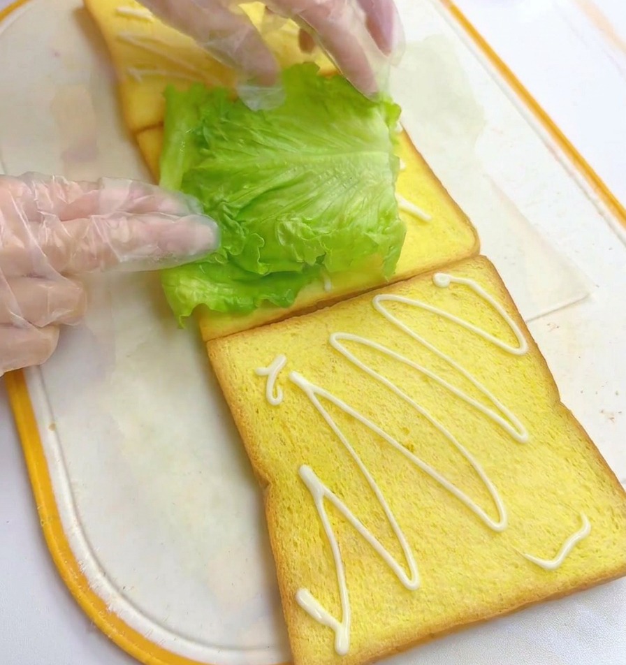 Hände platzieren ein frisches grünes Salatblatt auf eine Toastscheibe, daneben liegt eine weitere Scheibe mit Mayonnaise.