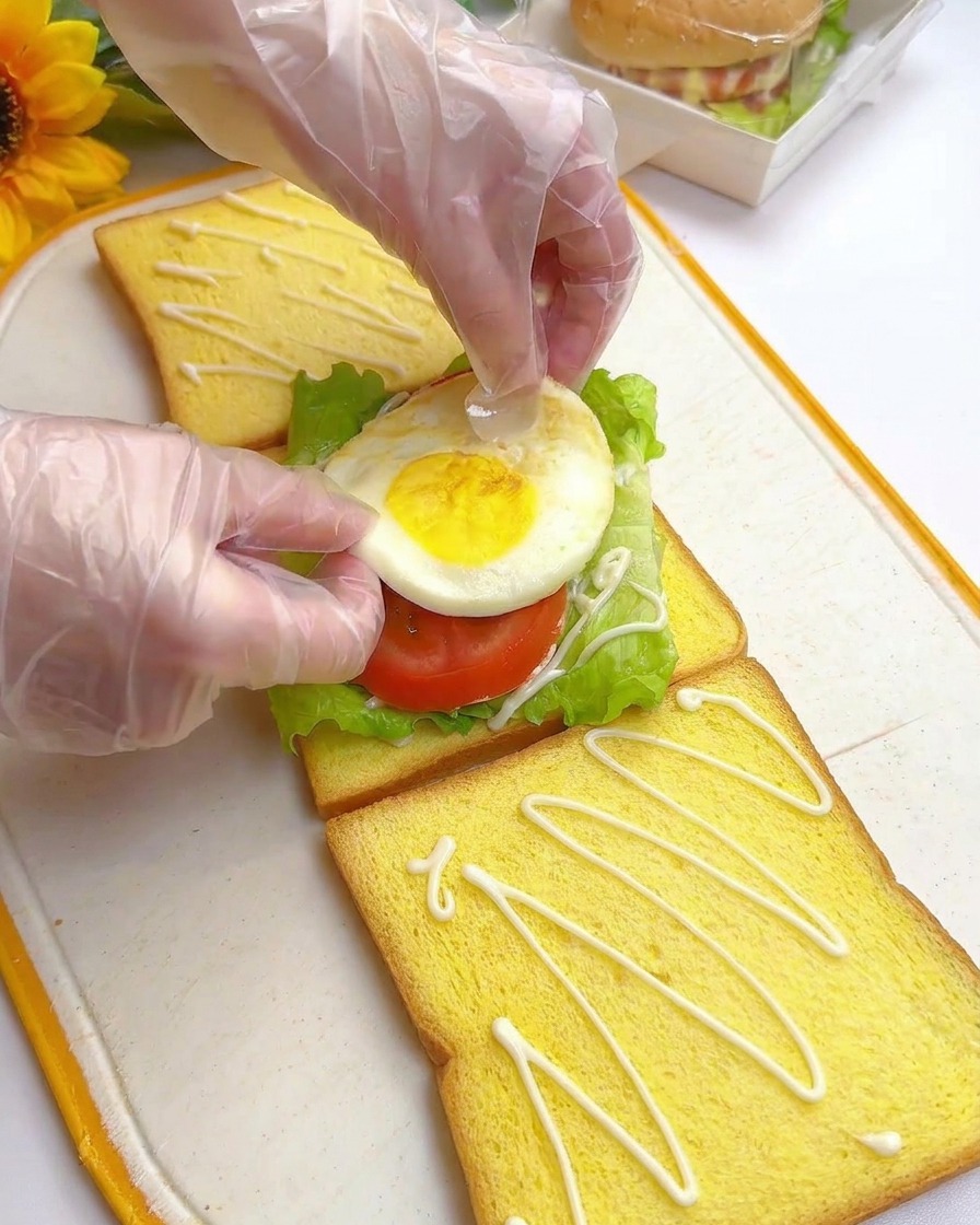 Hände platzieren vorsichtig ein gebratenes Ei auf Tomatenscheiben und Salat auf einer Toastscheibe.
