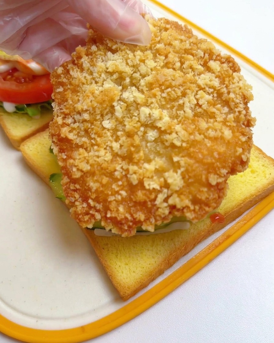 Eine Hand legt ein großes, goldbraun frittiertes Hähnchenschnitzel auf die Gurkenscheiben auf dem Toast.