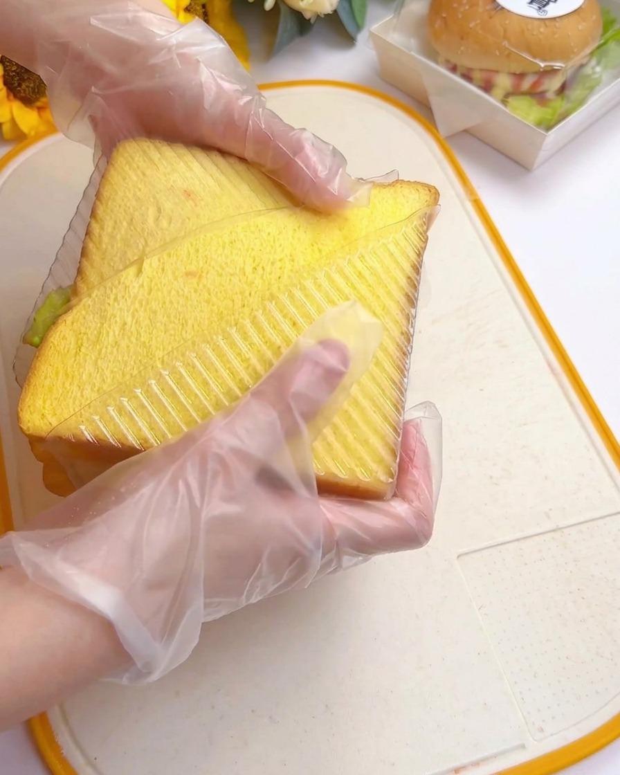 Hände drücken ein hohes Sandwich in eine durchsichtige, dreieckige Kunststoffform auf einem Schneidebrett.