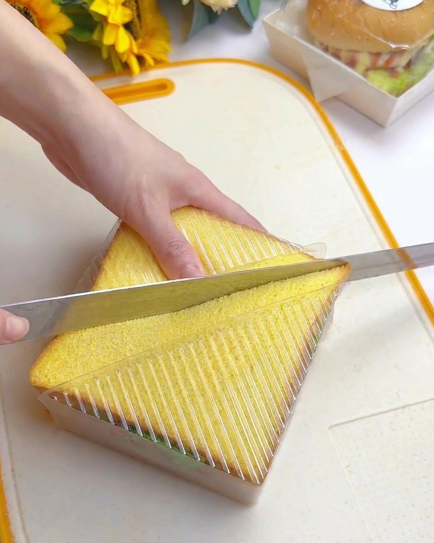 Eine Hand schneidet mit einem langen Wellenschliffmesser diagonal durch ein Sandwich, das in einer Kunststoffführung liegt.