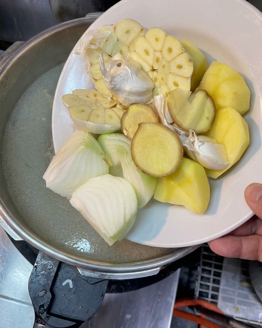 Ein Teller mit halbierten Knoblauchknollen, Zwiebeln, Ingwer und Kartoffelstücken über einem Topf mit kochender Brühe.