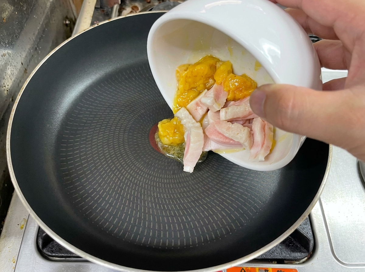 Eine Hand gibt Stücke von rohem Hühner- und Schweinefett aus einer weißen Schüssel in eine schwarze Pfanne.