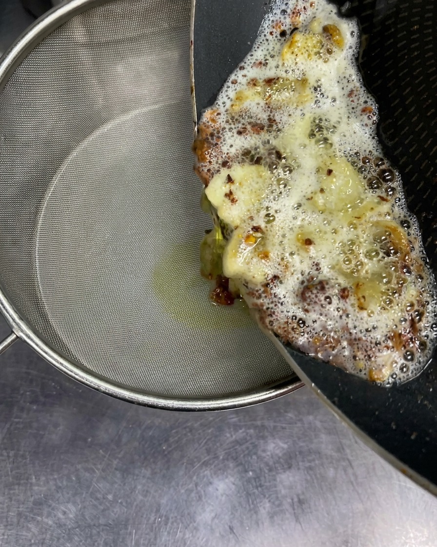 Heißes, sprudelndes Aroma-Öl mit frittierten Knoblauchstücken wird aus einer Pfanne in ein feines Sieb gegossen.