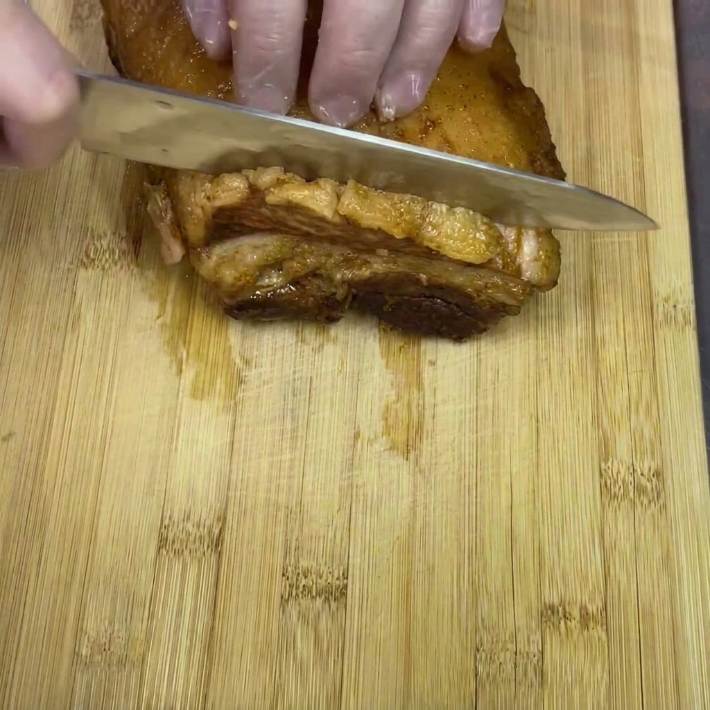 Hände mit Handschuhen schneiden ein Stück geschmorten Schweinebauch auf einem Holzbrett in dicke Scheiben.