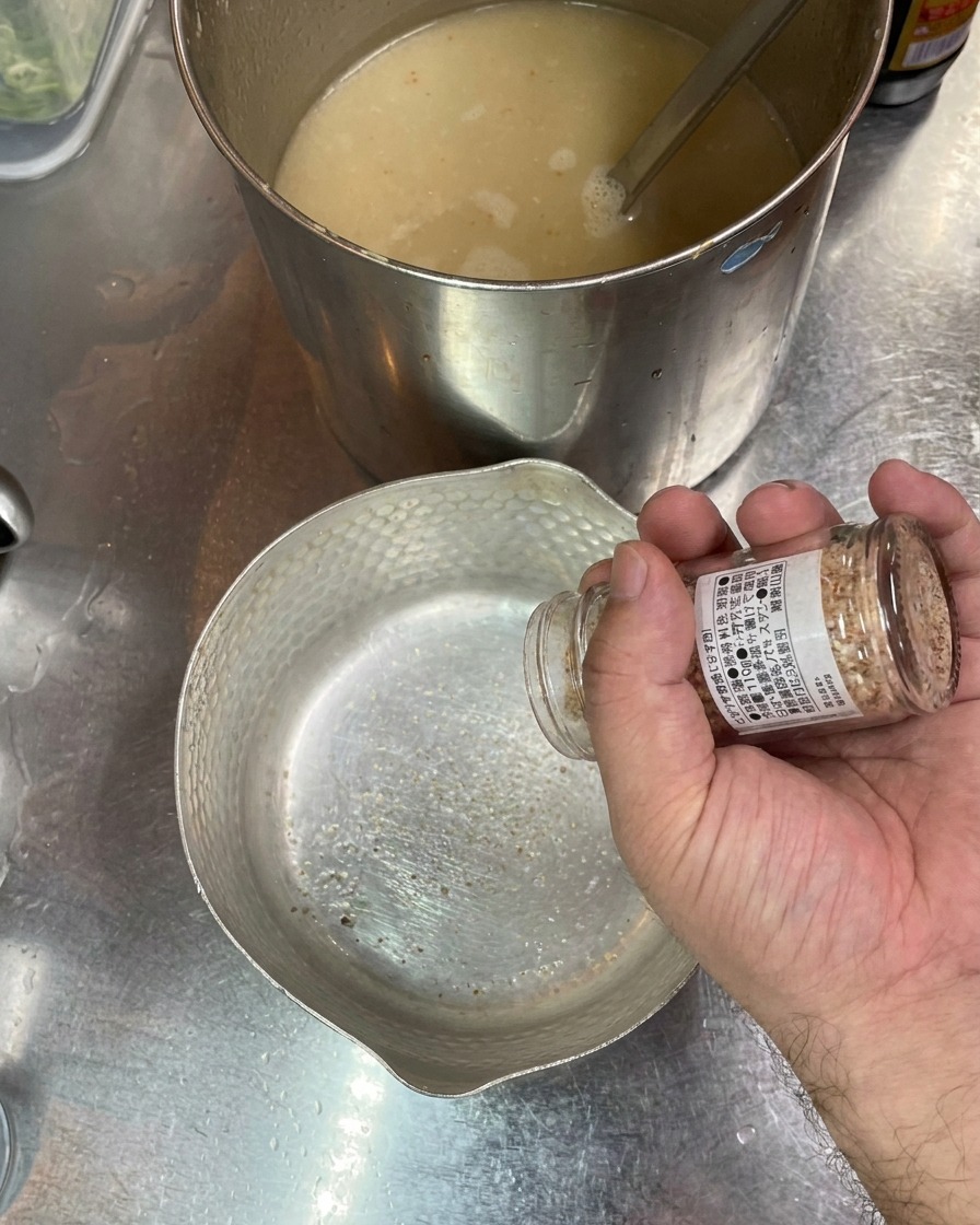 Eine Hand schüttelt Salz aus einem Glas in eine leere Metallschüssel, im Hintergrund steht ein Topf mit Brühe.
