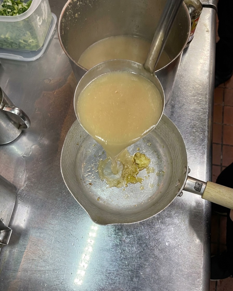 Eine Schöpfkelle gießt trübe, heiße Brühe in einen kleinen Topf, der Gewürze und Knoblauch enthält.