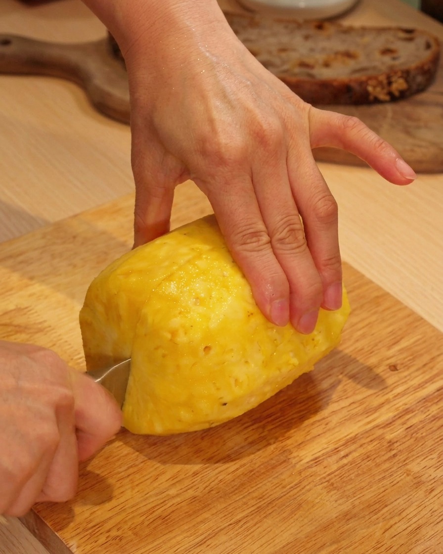 Hände schneiden eine geschälte frische Ananas auf einem Holzbrett in Scheiben.