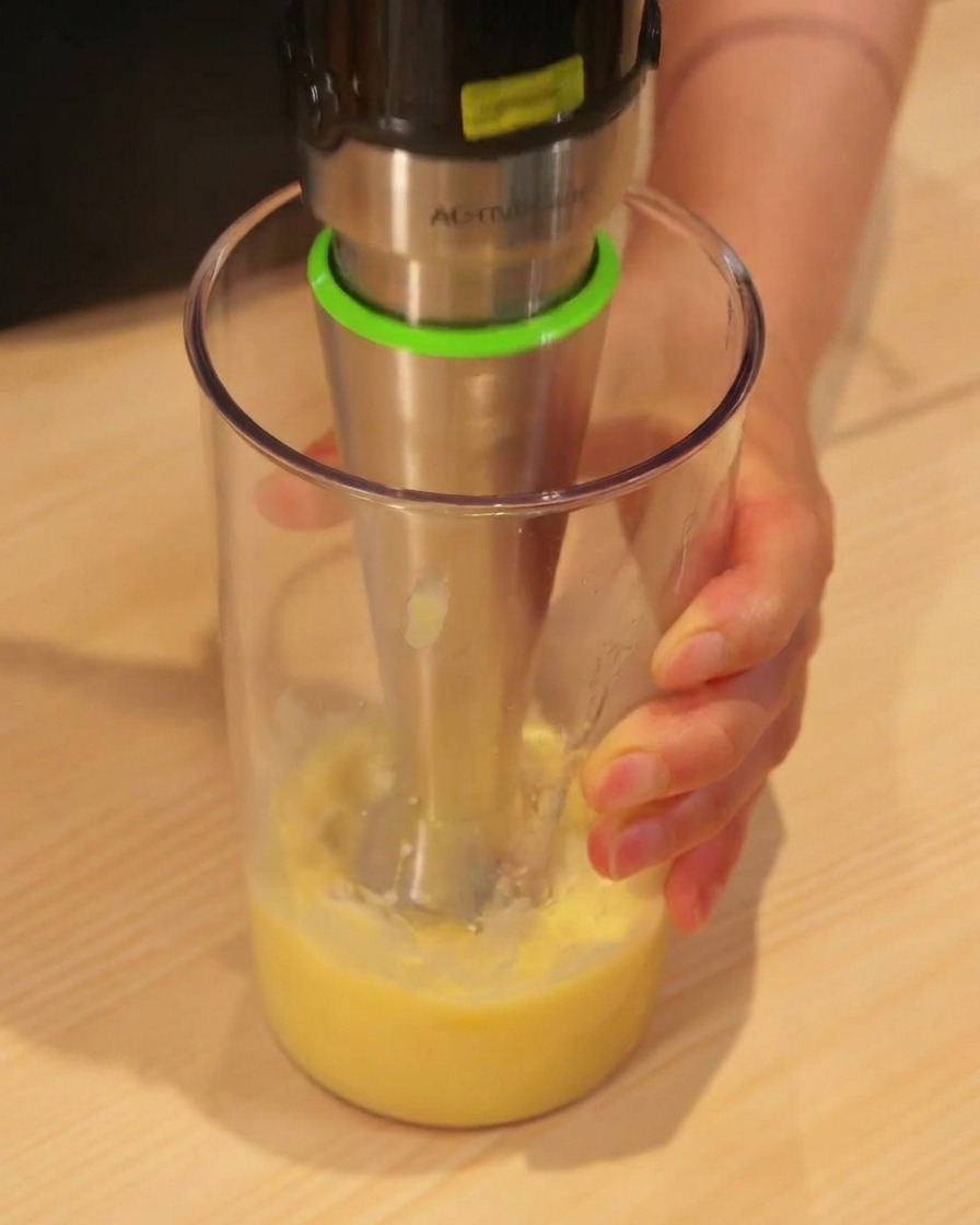 Ein Stabmixer püriert gelbe Ananasstücke in einem hohen Becher zu Saft.