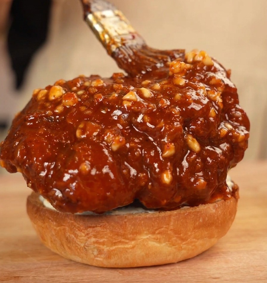 Ein großes, glasiertes Hähnchen-Patty liegt auf dem Brötchen und wird mit zusätzlicher Sauce bestrichen.