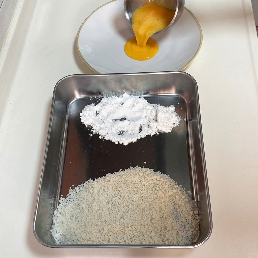 Verquirltes Ei wird in einen weißen Teller gegossen, daneben liegen Maisstärke und Panko auf einem Tablett.