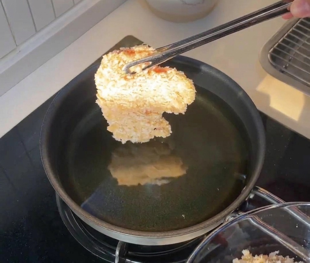 Ein paniertes Hähnchenfilet wird mit einer Zange über eine Pfanne mit heißem Öl gehalten.