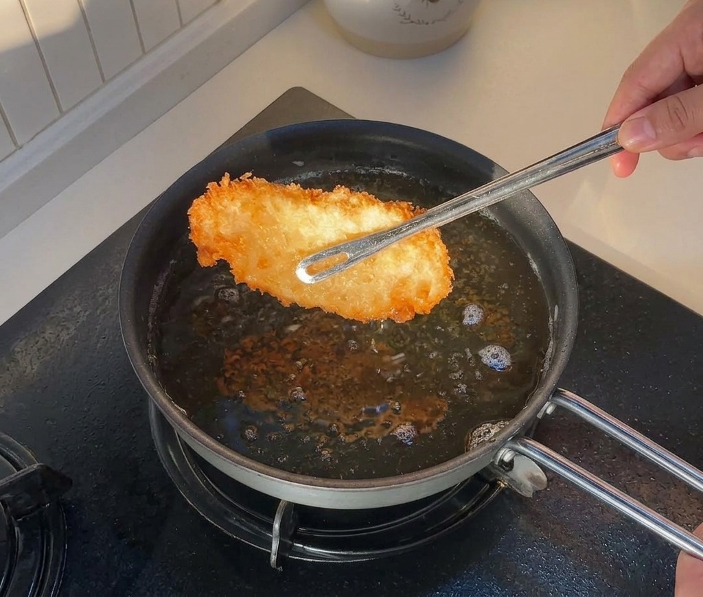 Eine Zange hebt ein goldbraun frittiertes Hähnchenstück aus einer Pfanne mit heißem Öl.
