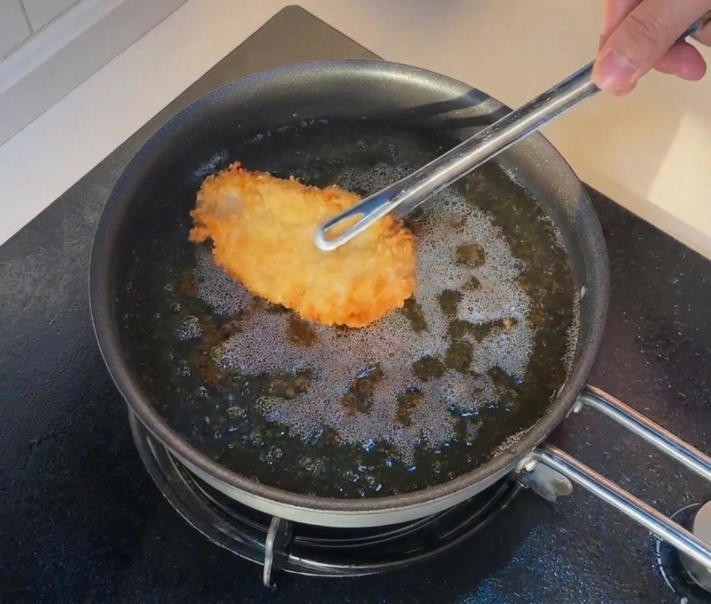 Eine Zange legt ein bereits frittiertes Hähnchenstück zurück in sprudelnd heißes Öl.