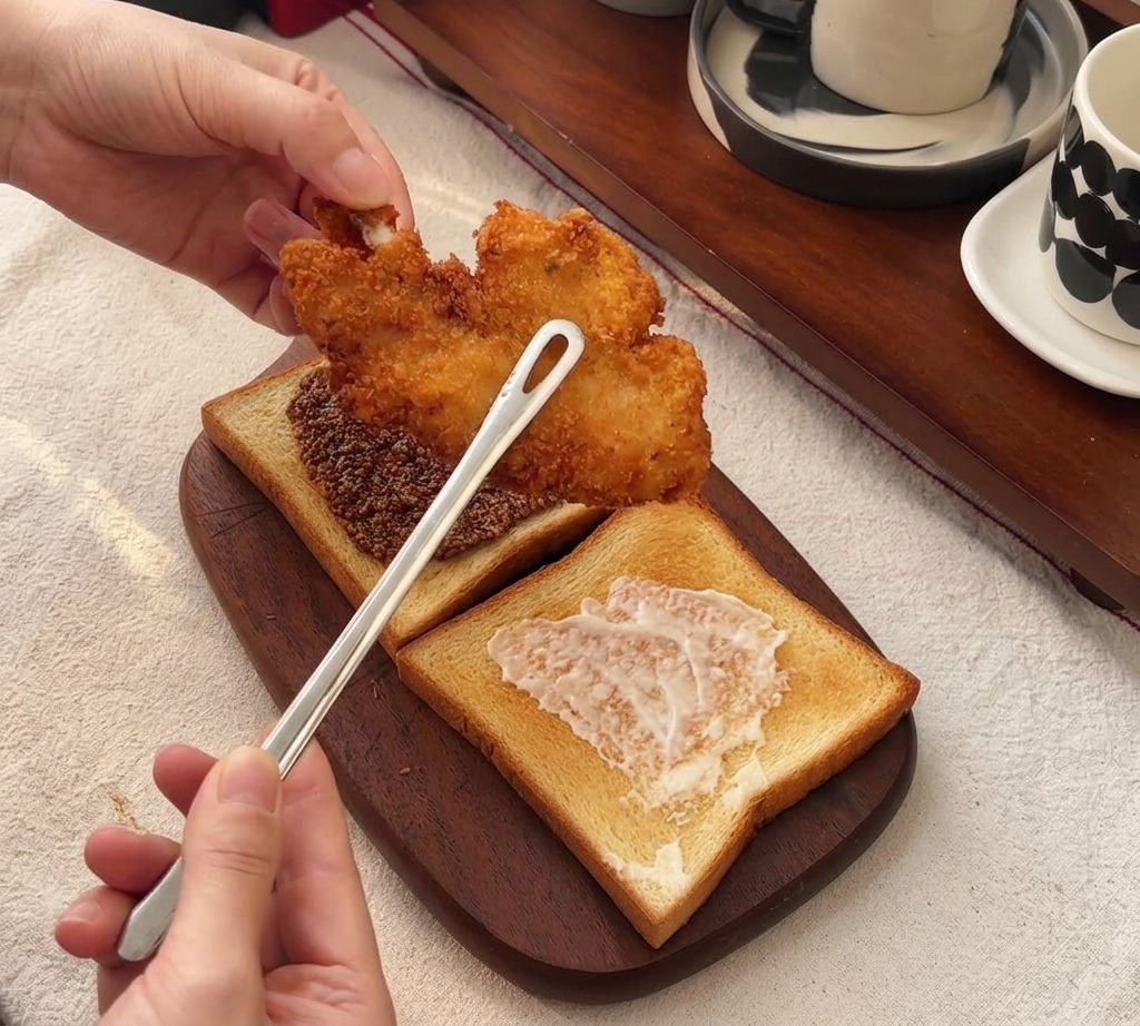Eine Zange legt ein großes, knusprig frittiertes Hähnchenfilet auf einen Toast mit Honig-Senf.