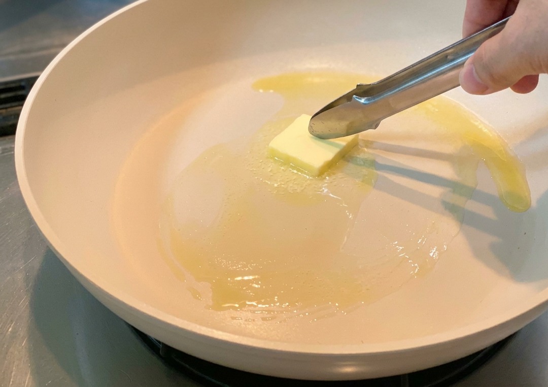 Ein Stück Butter schmilzt in einer hellen Pfanne und wird mit einer Zange verteilt.