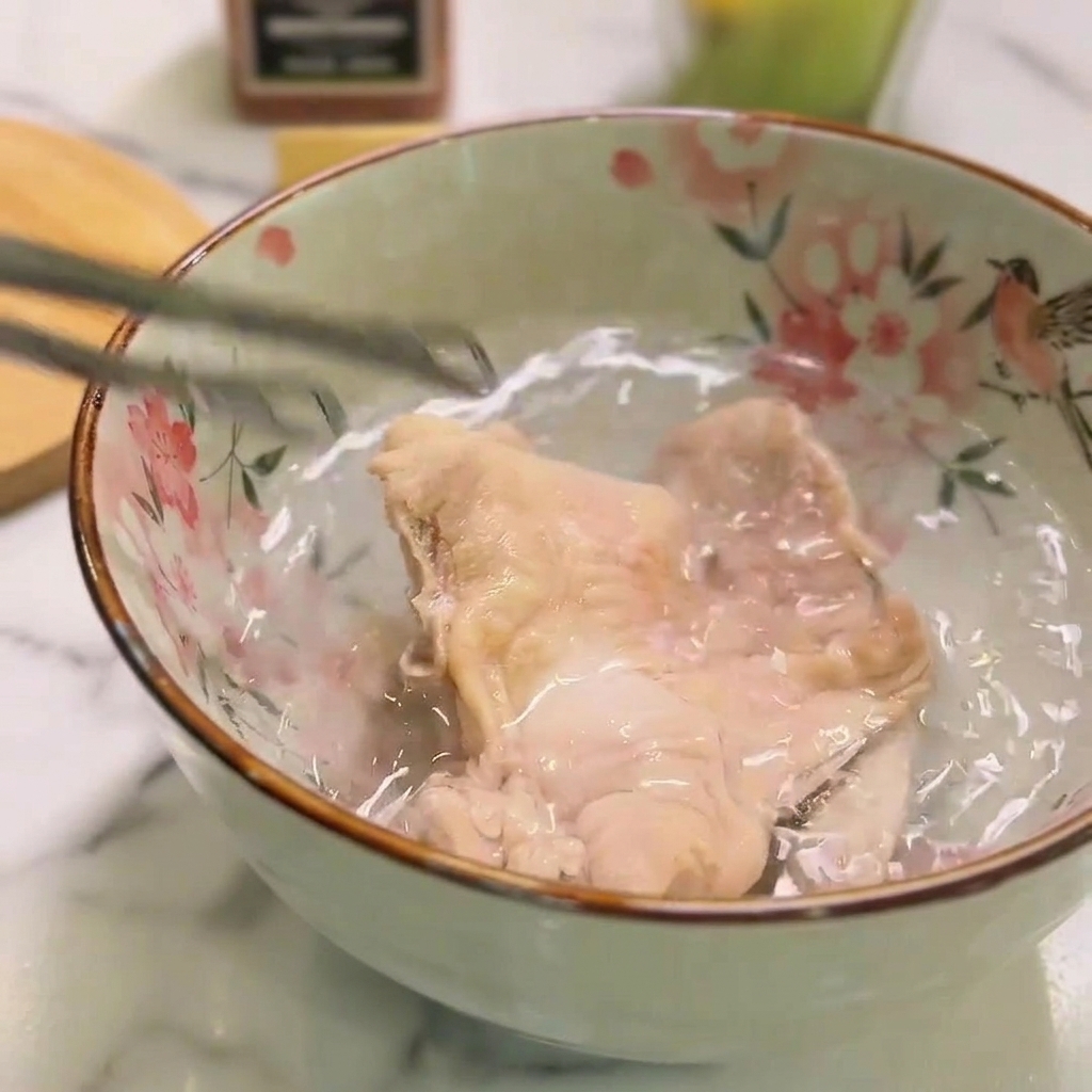 Ein Stück gekochtes Hähnchen liegt in einer Keramikschüssel mit kaltem Wasser.
