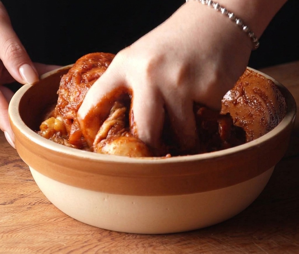 Eine Hand mischt rohe Hähnchenschenkel, die mit einer dunkelroten Marinade überzogen sind.