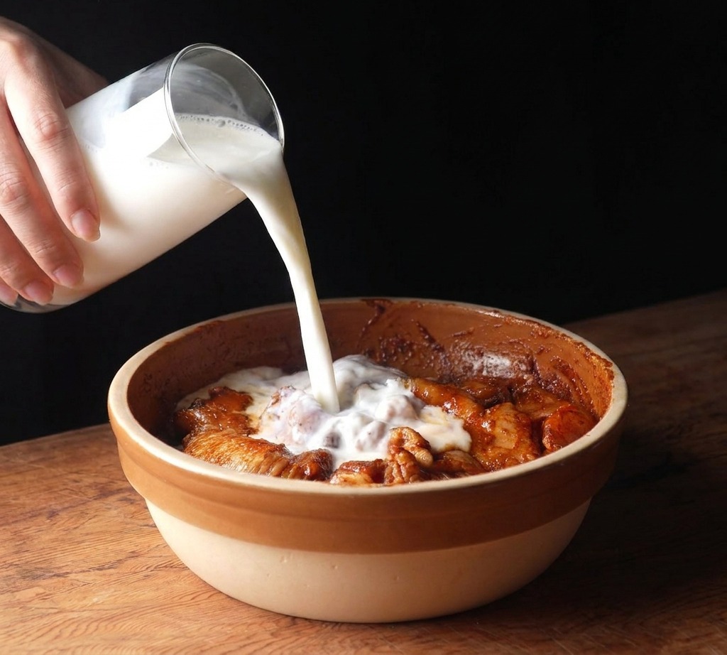 Buttermilch wird aus einem Glaskrug in eine Schüssel mit mariniertem Hähnchen gegossen.