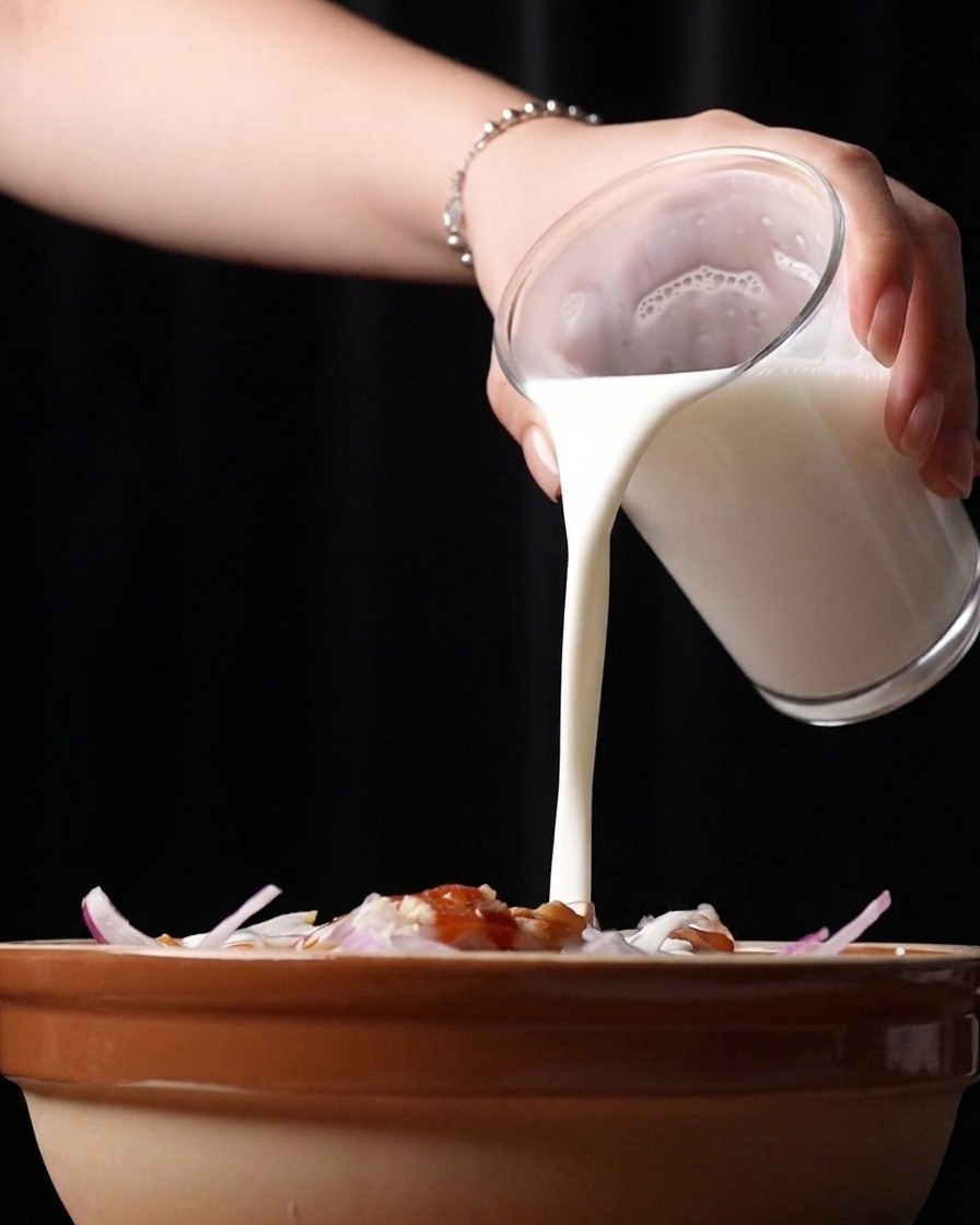 Weiße Buttermilch wird aus einem Glas über das marinierte Hähnchen gegossen.