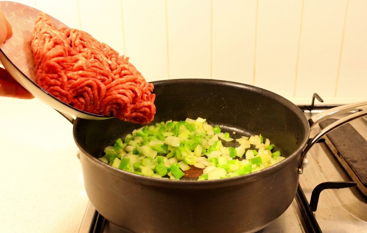 Frisches rohes Rinderhackfleisch wird von einem Teller in eine Pfanne mit gedünstetem Gemüse gegeben.