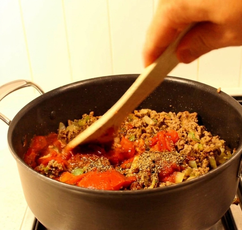 Ein Holzspatel vermischt Hackfleisch, Paprika und eine rote Tomatensauce in einem schwarzen Kochtopf.