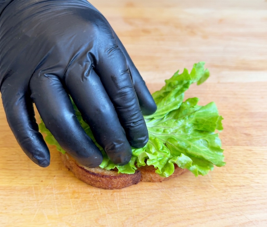 Eine Hand legt ein gefaltetes Blatt grünen Salat auf eine geröstete Brotscheibe auf einem Holzbrett.