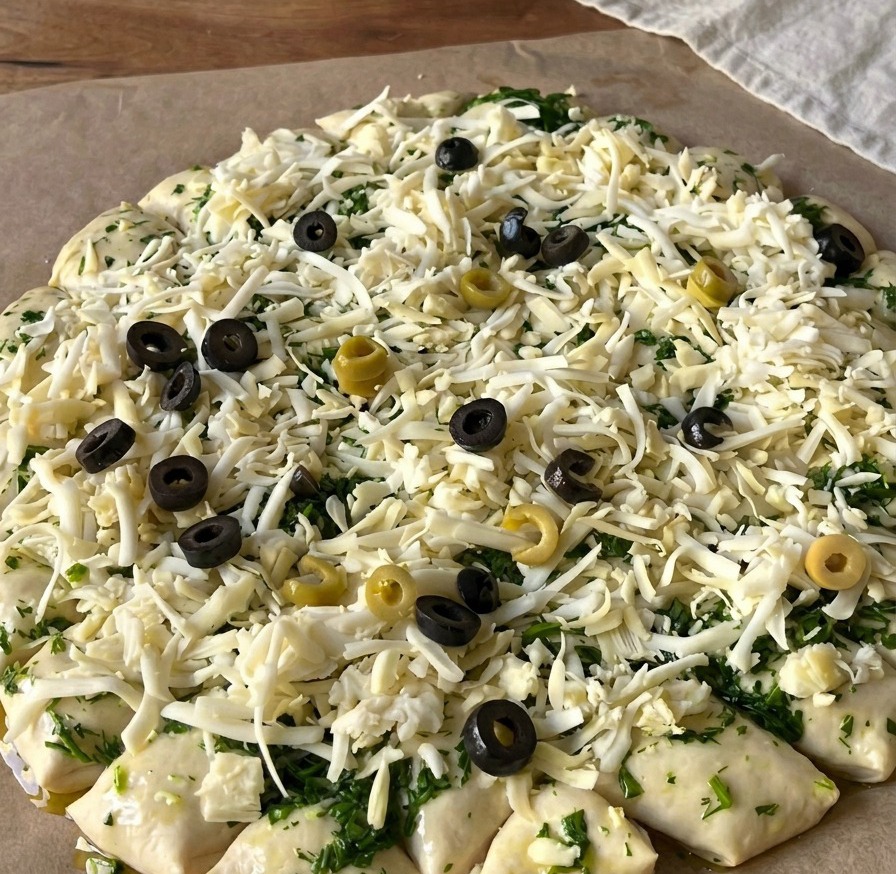 Draufsicht auf den rohen Pizzateig, der mit Käse bedeckt und mit zahlreichen schwarzen und grünen Olivenscheiben belegt ist.