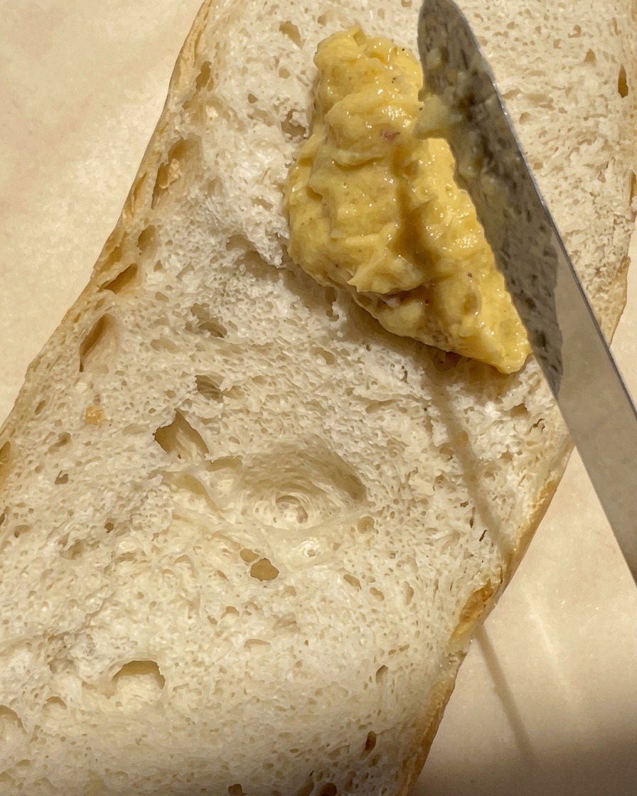 Nahaufnahme eines Messers, das eine dicke, cremige Mischung aus geröstetem Knoblauch und Butter auf die luftige Oberfläche eines halbierten Baguettes streicht.