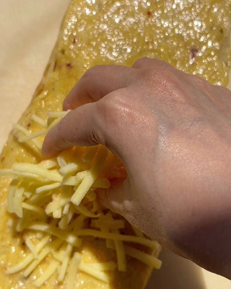 Eine Hand streut eine große Handvoll geriebenen weißen Mozzarella auf ein mit Knoblauchbutter bestrichenes Brot.