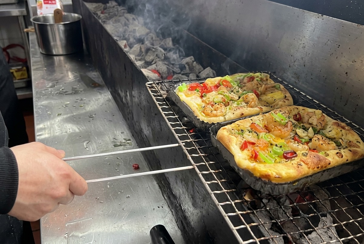 Eine mit Garnelen und buntem Gemüse belegte, rechteckige Pizza wird in einer Profiküche auf einen langen Holzkohlegrillrost gelegt.