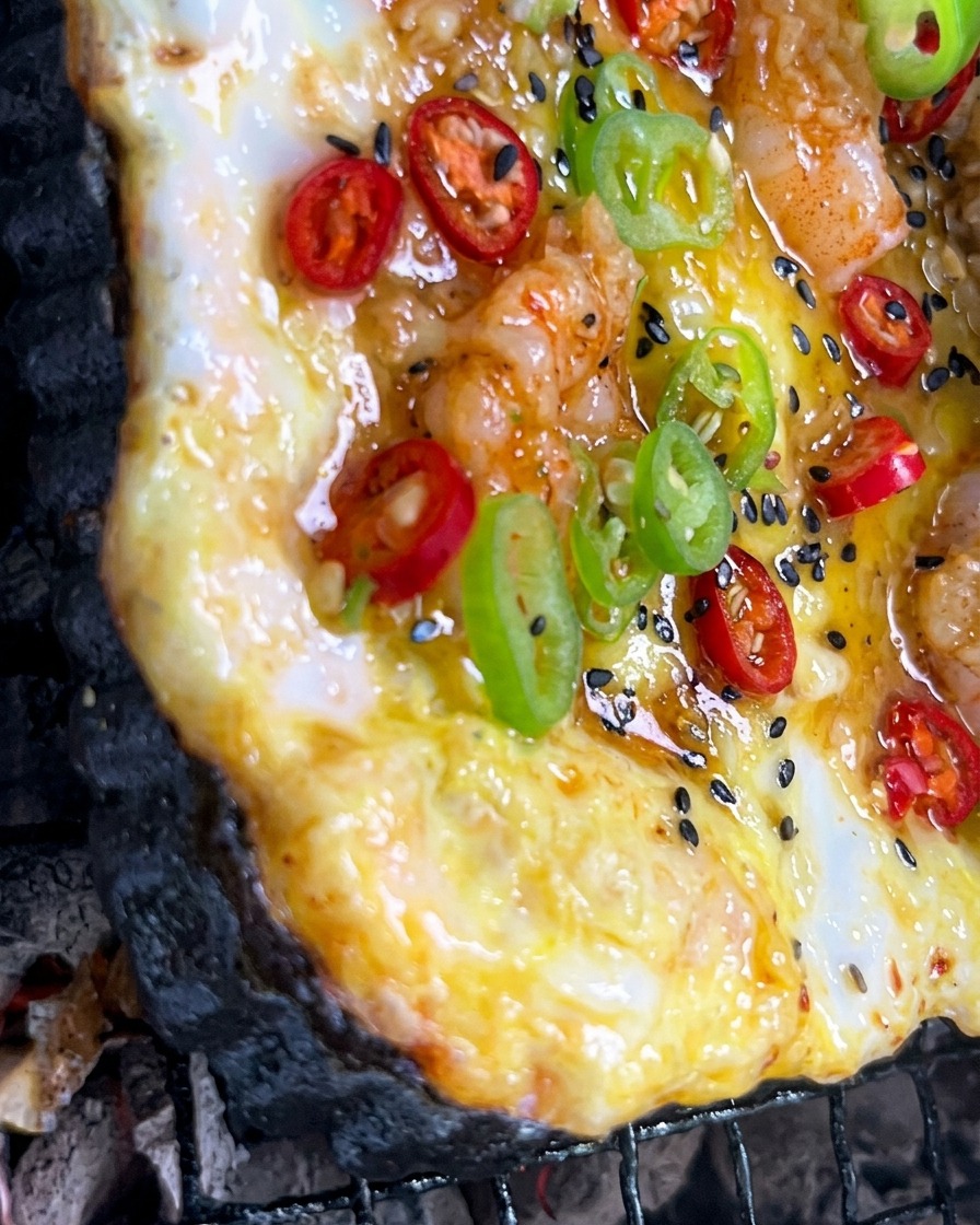 Gehackte Knoblauchsauce wird mit einem Pinsel auf eine bunte Pizza gestrichen, die über Holzkohle grillt.