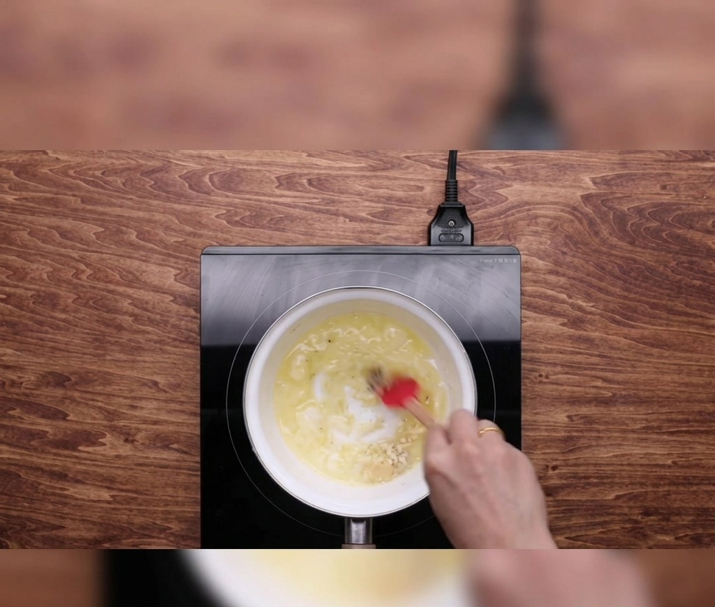Eine Hand rührt geschmolzene Butter und gehackten Knoblauch mit einem roten Teigschaber in einem kleinen weißen Topf auf einem schwarzen Induktionskochfeld.