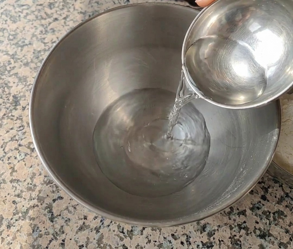 Wasser wird aus einer kleinen Metallschüssel in eine große Rührschüssel aus Edelstahl auf einer Granitarbeitsplatte gegossen.