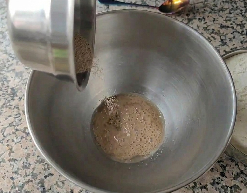 Granulierte Trockenhefe wird in eine Edelstahlschüssel mit Wasser gegeben, auf dessen Oberfläche sich kleine Bläschen bilden.