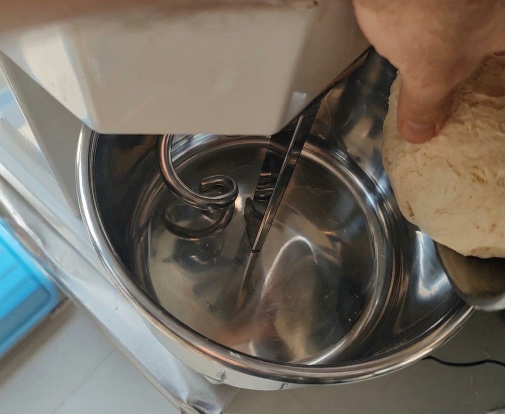 Eine Hand gibt ein Stück des fermentierten, texturierten Biga-Teigs in eine große Rührschüssel einer Knetmaschine.