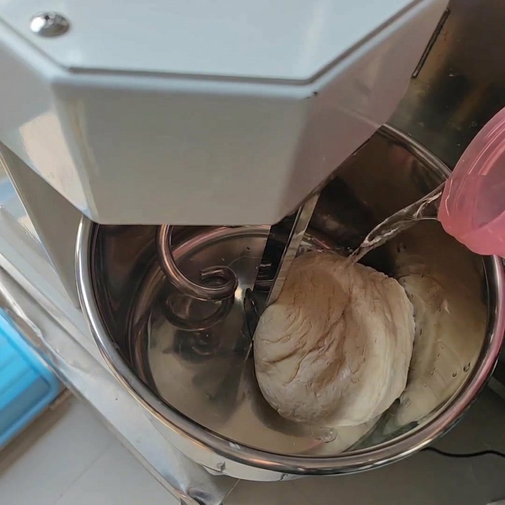 Wasser wird aus einem rosafarbenen Behälter in eine Edelstahl-Knetschüssel gegossen, in der sich bereits ein Teig befindet.