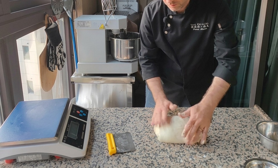 Eine große Menge heller Pizzateig wird mit einem gelben Teigabstecher auf einer gesprenkelten Granitarbeitsplatte neben einer Digitalwaage portioniert.