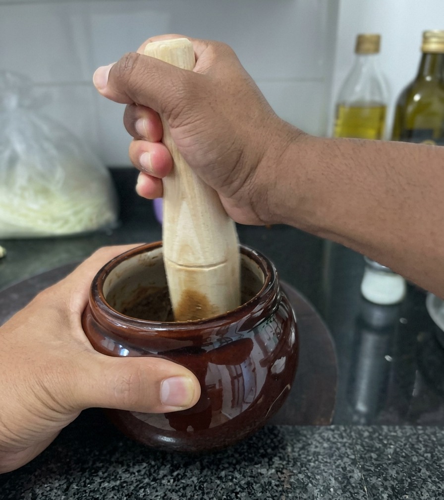 Eine Person zerbricht dunklen, frittierten Knoblauch mit einem hölzernen Stößel in einem kleinen braunen Keramikmörser auf einer Granitarbeitsplatte.