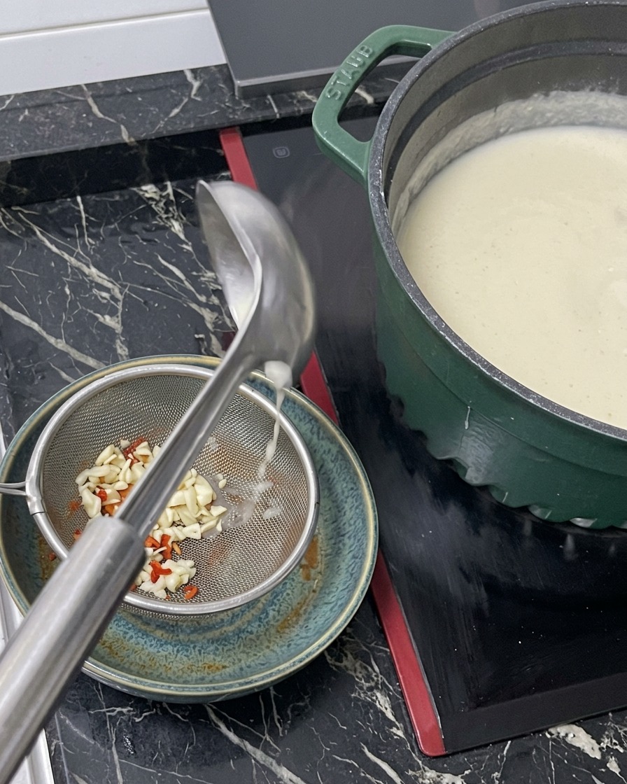 Heiße weiße Brühe wird aus einer Kelle durch ein Metallsieb in eine Schale gegossen, die bereits die Ramen-Würzbasis enthält.