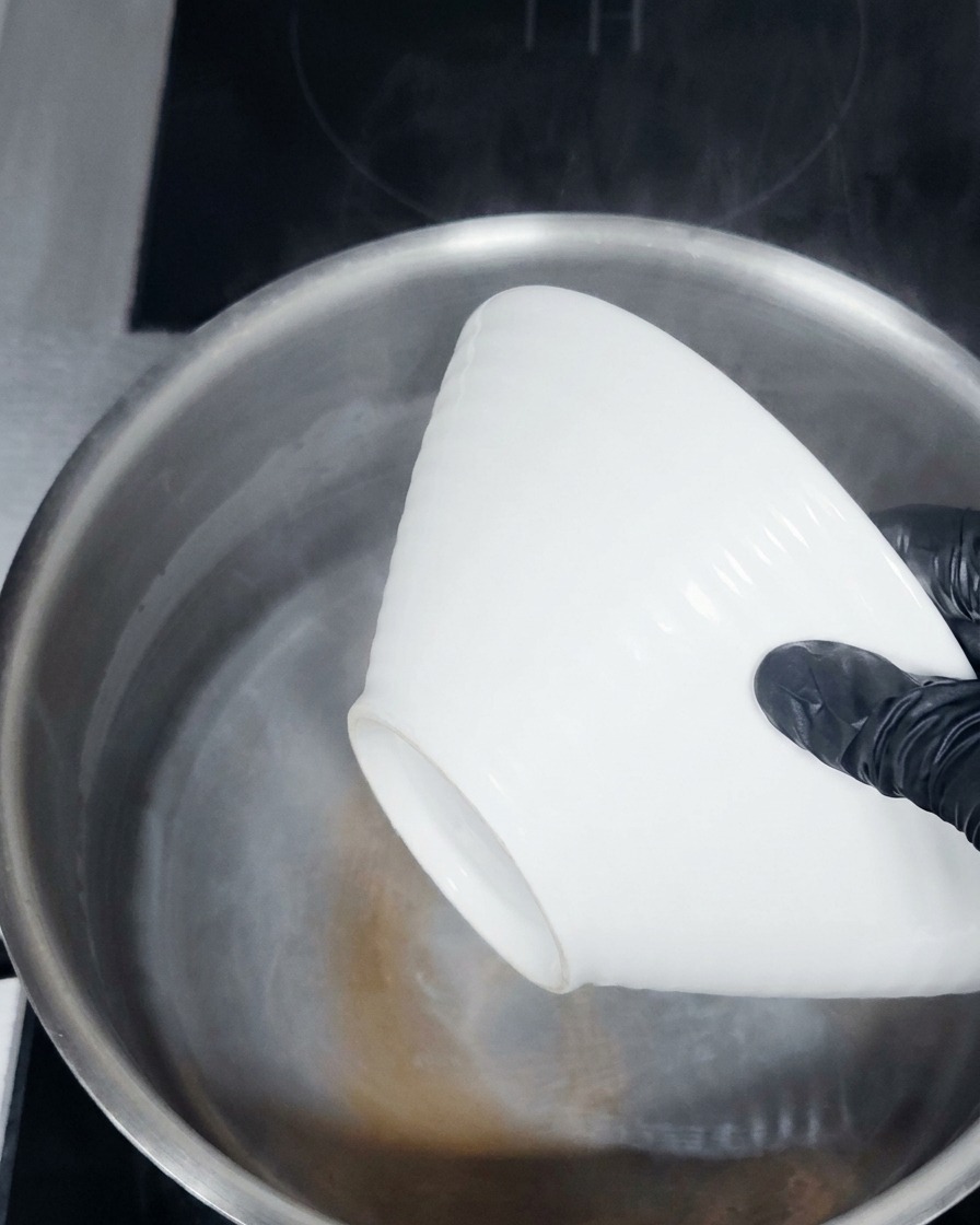 Hände in Handschuhen senken eine weiße Keramikschüssel in einen Topf mit dampfend heißem Wasser.