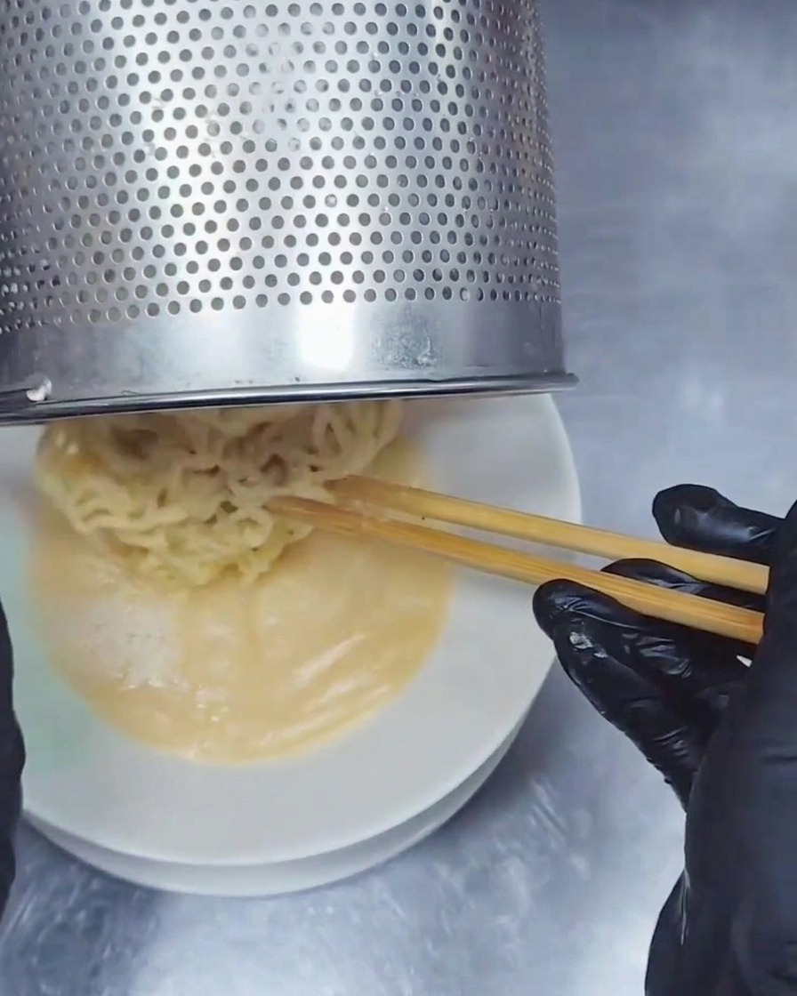 Ein Nudelsieb aus Edelstahl gibt wellige Ramen-Nudeln in eine Schüssel mit cremiger Tonkotsu-Brühe.